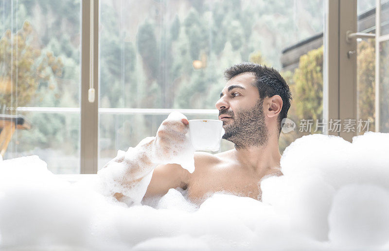 一个男人在热水浴缸里，拿着一杯茶/咖啡，看着外面